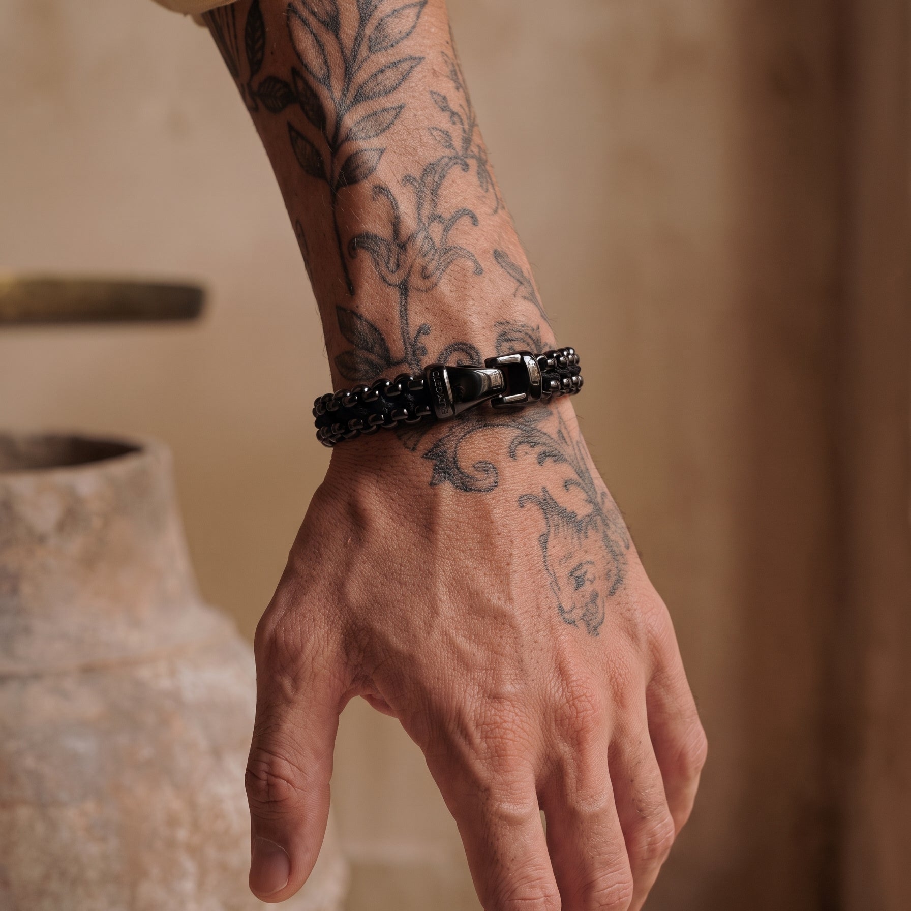 A close-up of a tattooed hand and wrist wearing a black beaded bracelet with a silver-toned rectangular clasp, set against a warm, rustic background with a blurred clay pot.