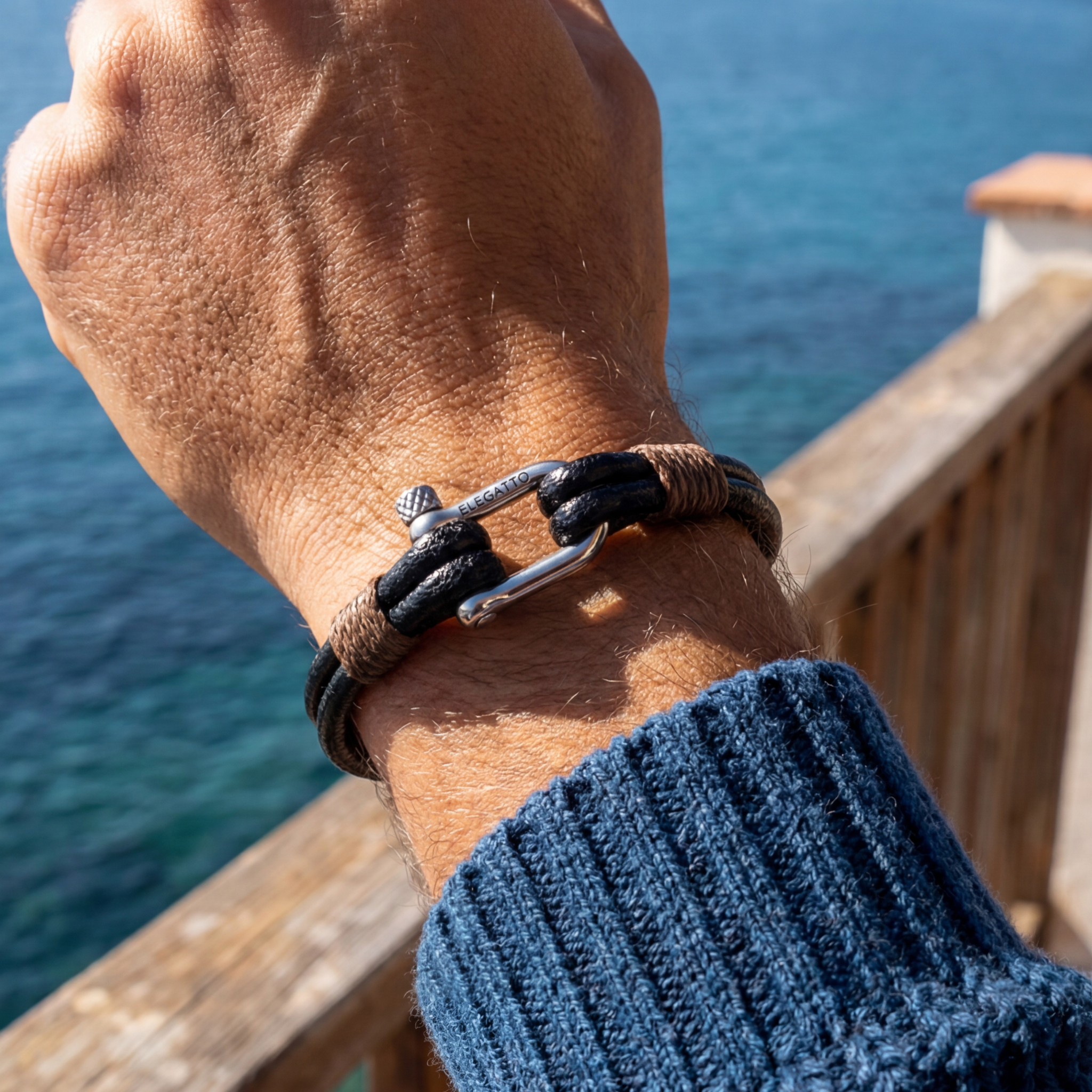 Hand wearing a leather bracelet with a blue sweater sleeve, overlooking water.