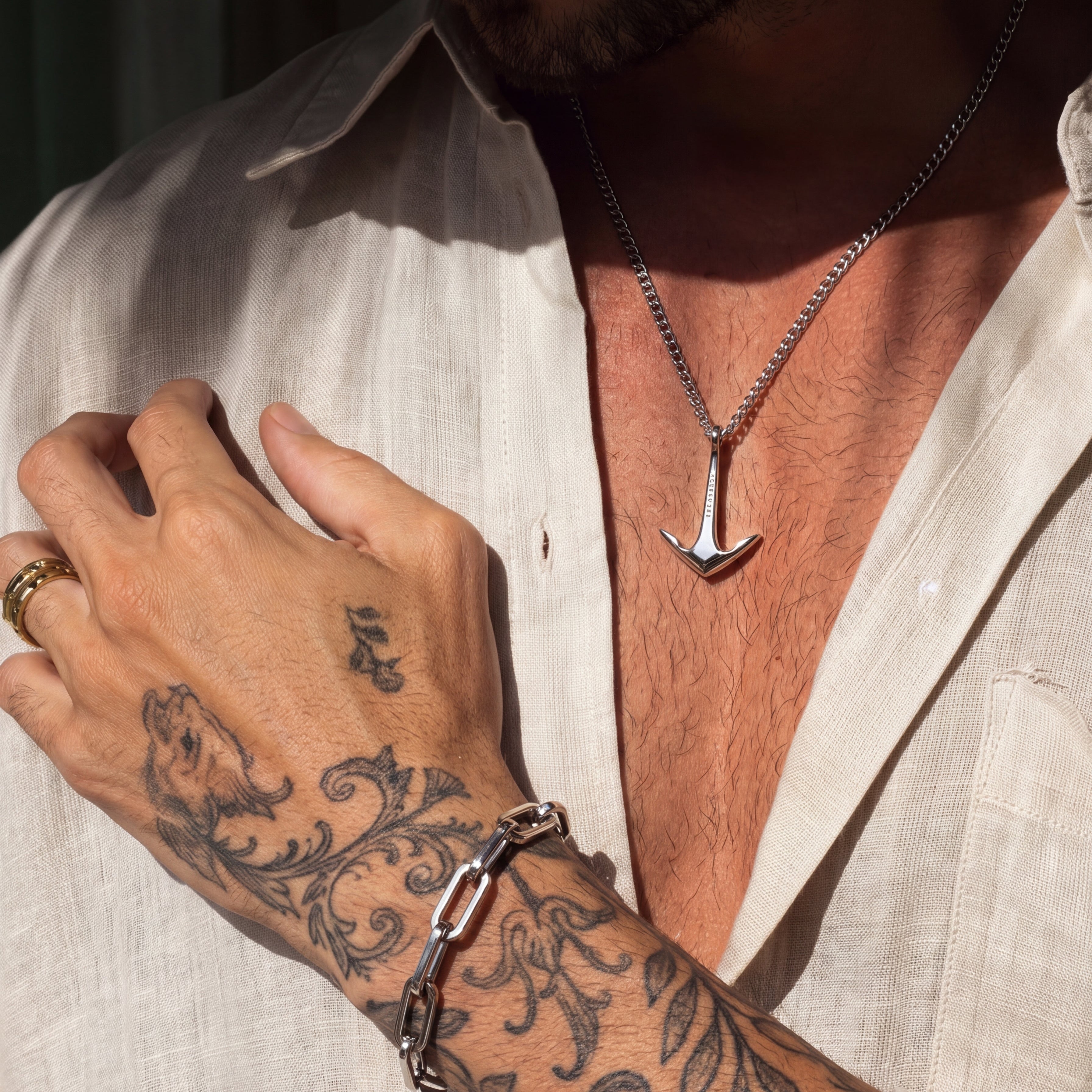 Man wearing a silver anchor necklace and bracelet with tattoos on his arm.