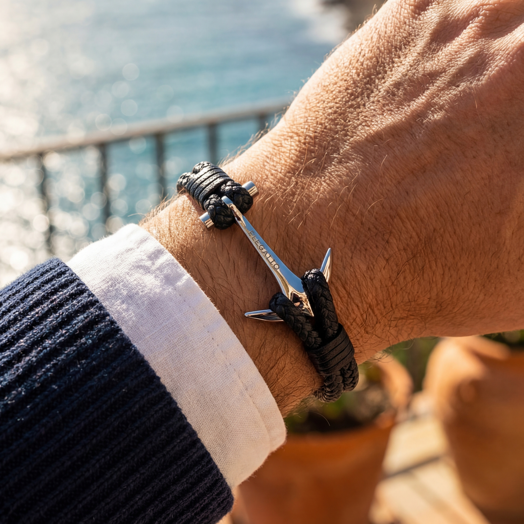 Hand wearing a bracelet with an anchor charm against a blurred coastal background