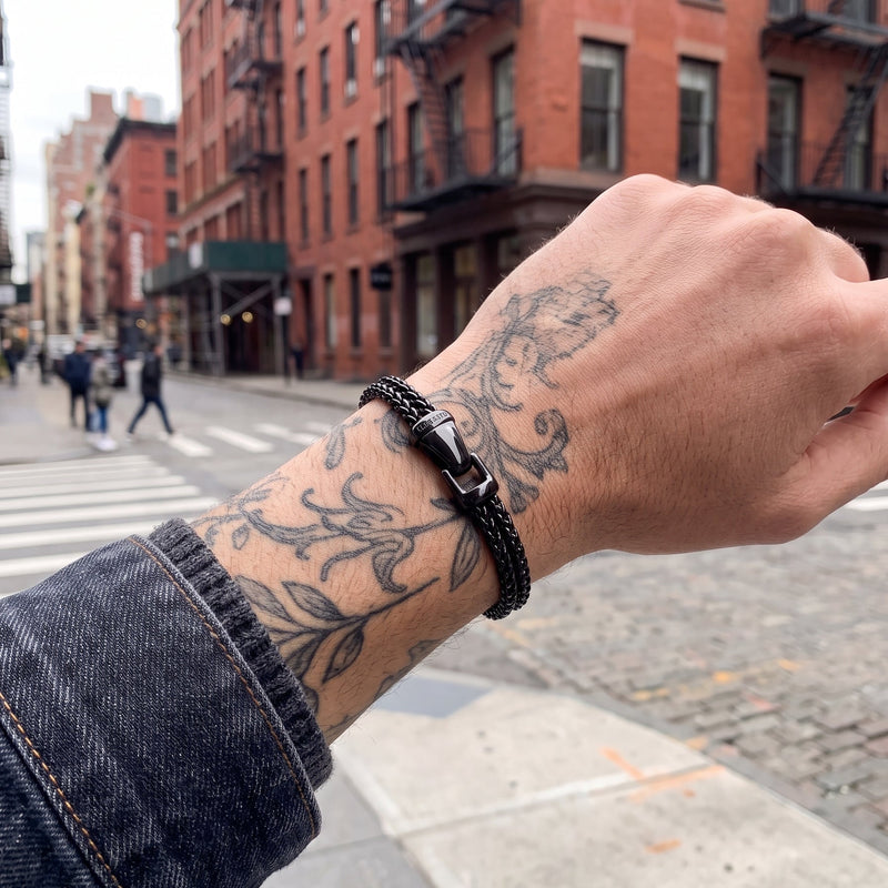 A close-up of a tattooed wrist wearing a black braided cord bracelet with a silver-toned metal clasp, set against a blurred city street background with brick buildings.